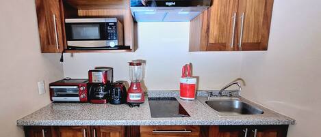 A compact kitchen with wooden cabinets, a microwave, a toaster, a coffee maker, a blender, and a sink.