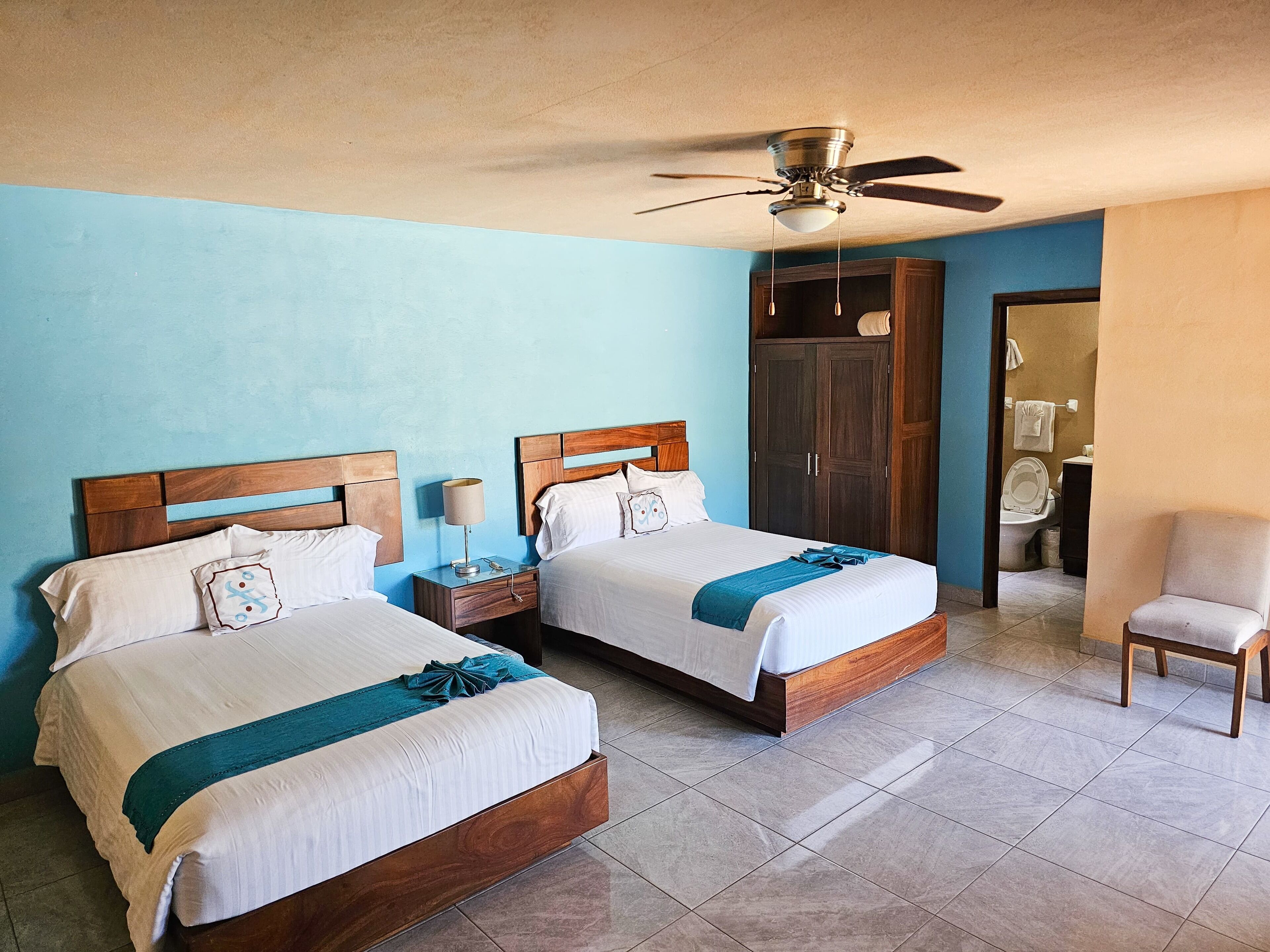 Habitación de hotel con dos camas, ventilador de techo, armario y baño visible a través de una puerta abierta.