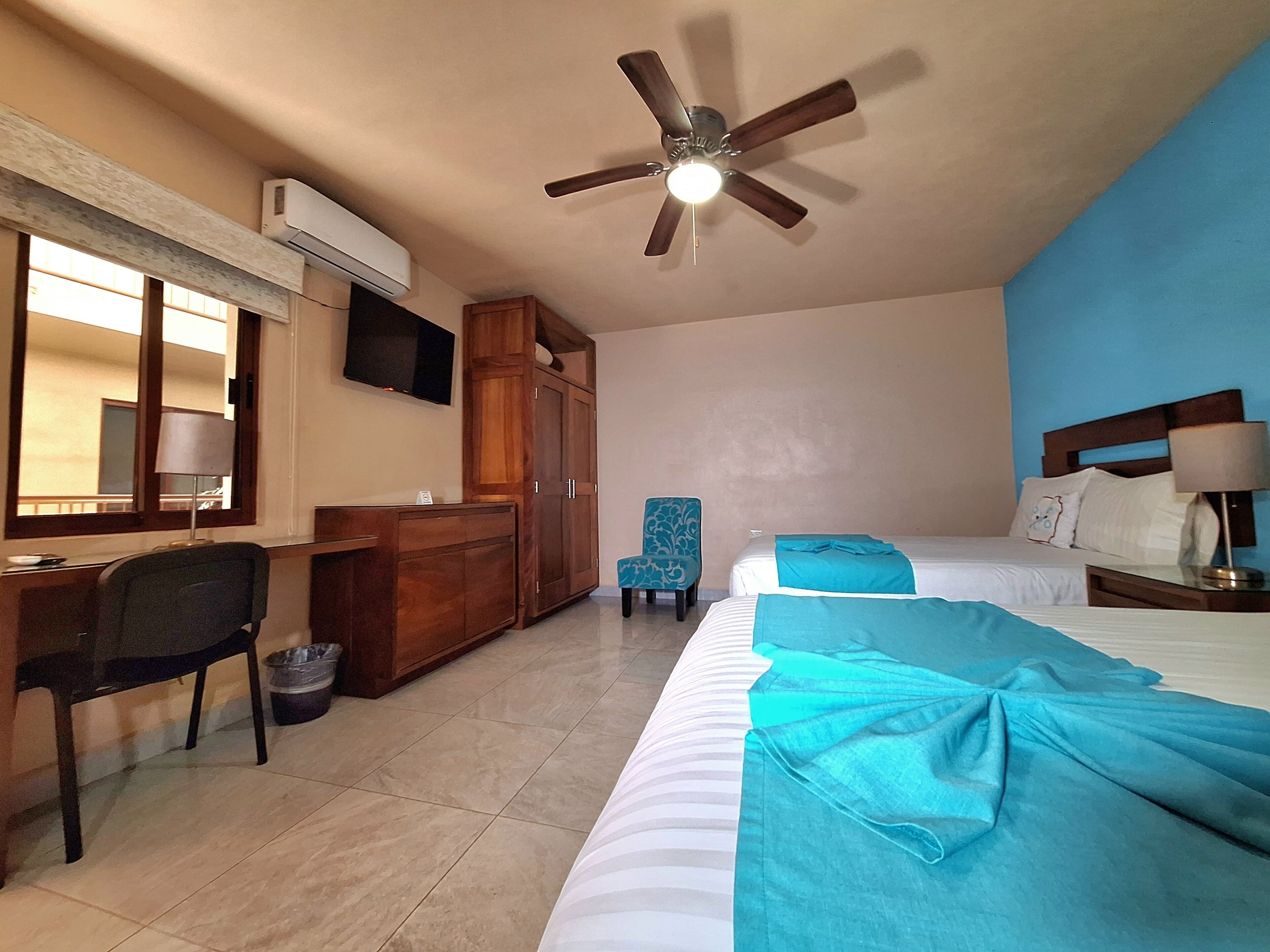 A hotel room with a bed, a desk with a chair, a wardrobe, a ceiling fan, and a TV mounted on the wall.