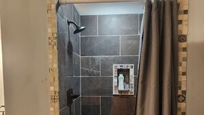 A shower area with dark tiles, a showerhead, and a small shelf with toiletries.