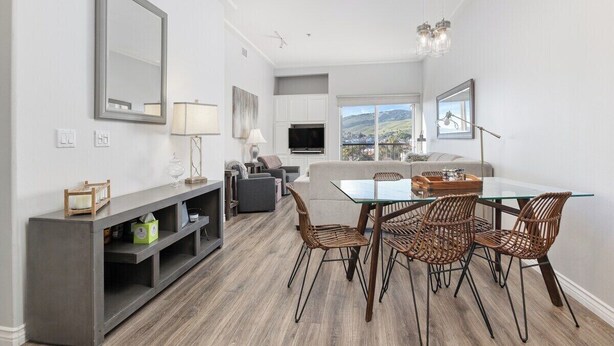 A modern living room with a glass dining table, wicker chairs, a grey sofa, and a TV mounted on the wall.