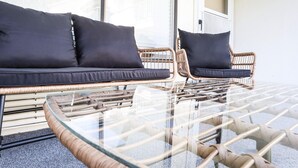 A wicker sofa with dark cushions and a glass-top table in a bright room with a large window.