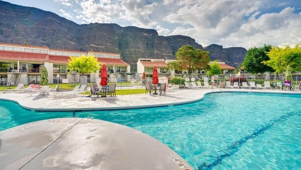 A swimming pool with lounge chairs and umbrellas, surrounded by buildings with red roofs and a mountainous backdrop.