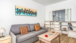 Una sala de estar moderna con un sofá gris, dos cojines estampados, una mesa de centro de madera y una estantería con libros y objetos decorativos.
