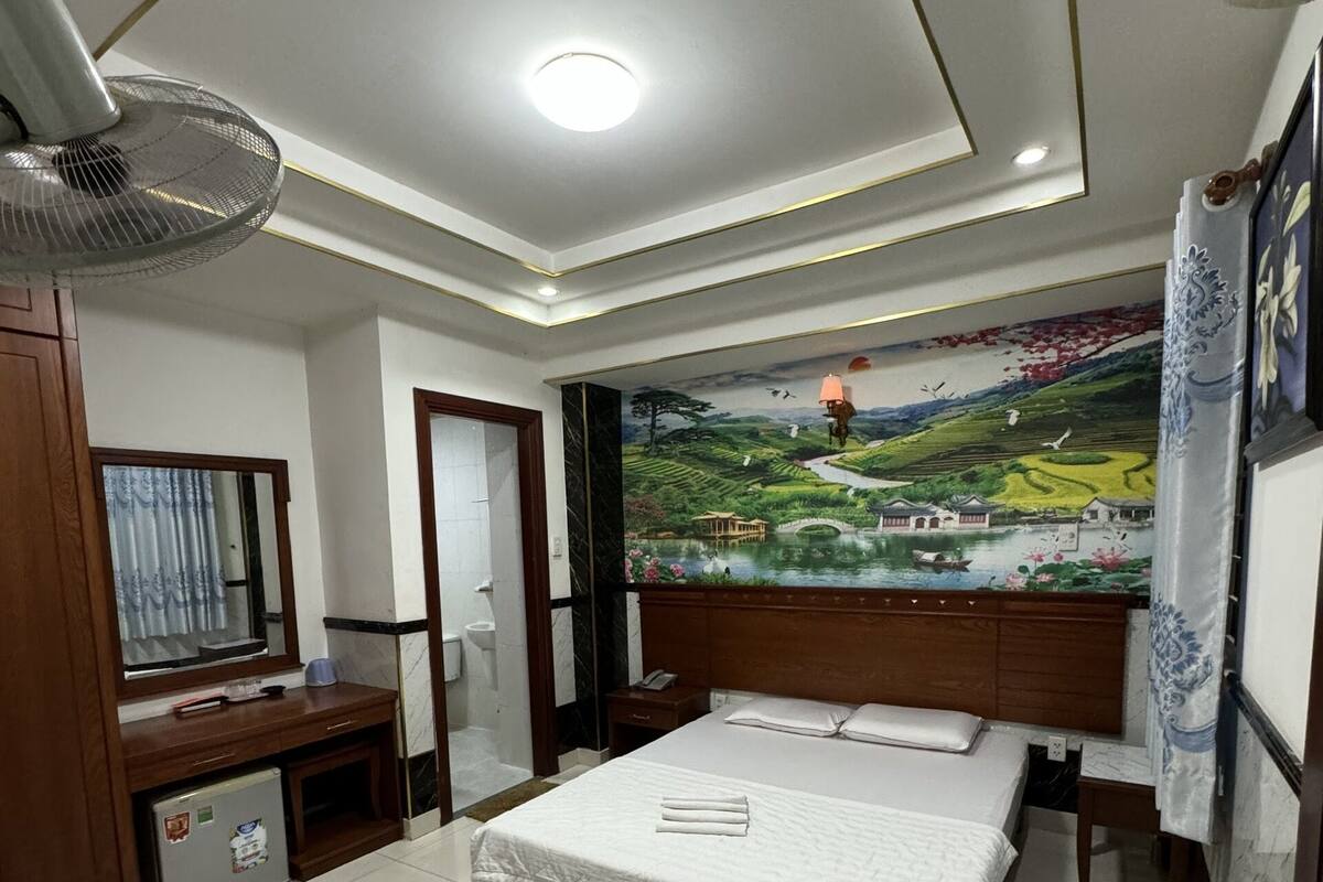 Ein Hotelzimmer mit einem großen Bett, einem Schreibtisch mit Stuhl, einem kleinen Kühlschrank, einem Standventilator und einem Wandgemälde.