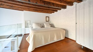 Une chambre avec un plafond en bois, un mur en briques blanches, un lit recouvert d’une courtepointe blanche et une grande armoire.