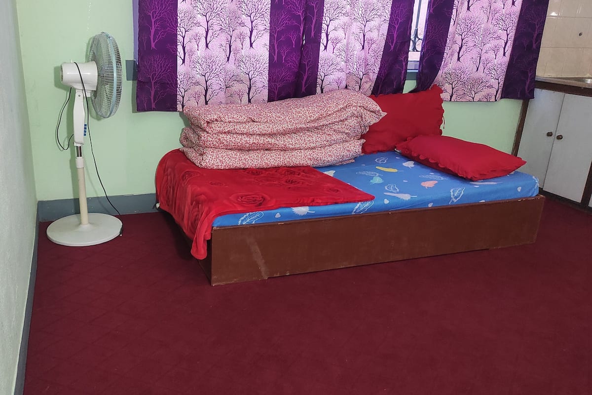 A single bed with a red and blue floral bedspread, a red blanket, and two red pillows. A standing fan is against the wall.