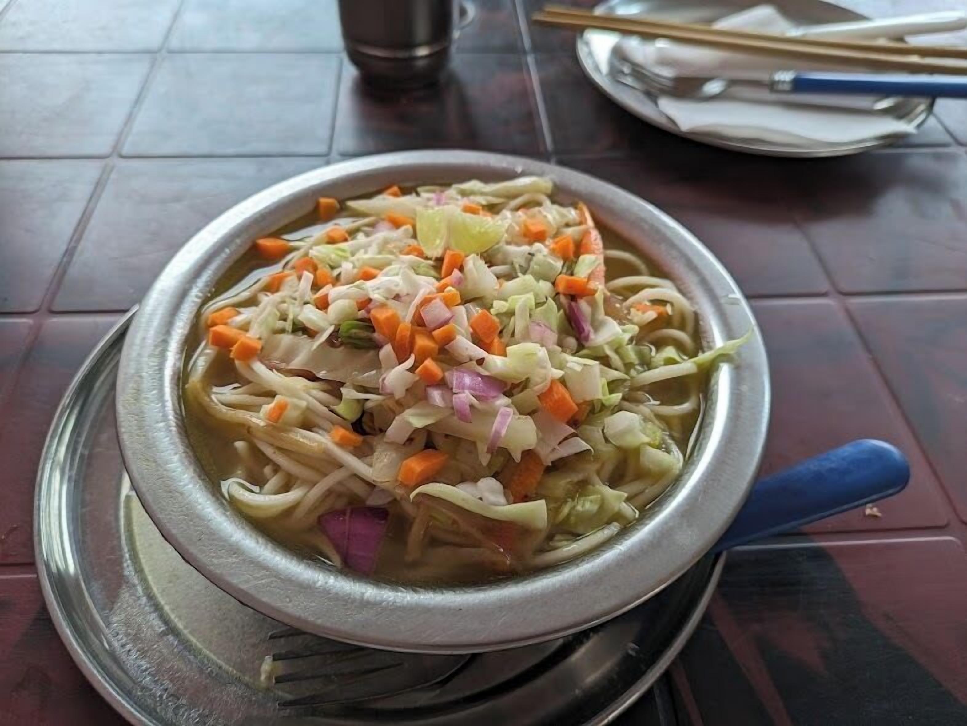 A bowl of noodle soup with vegetables on a metal plate.