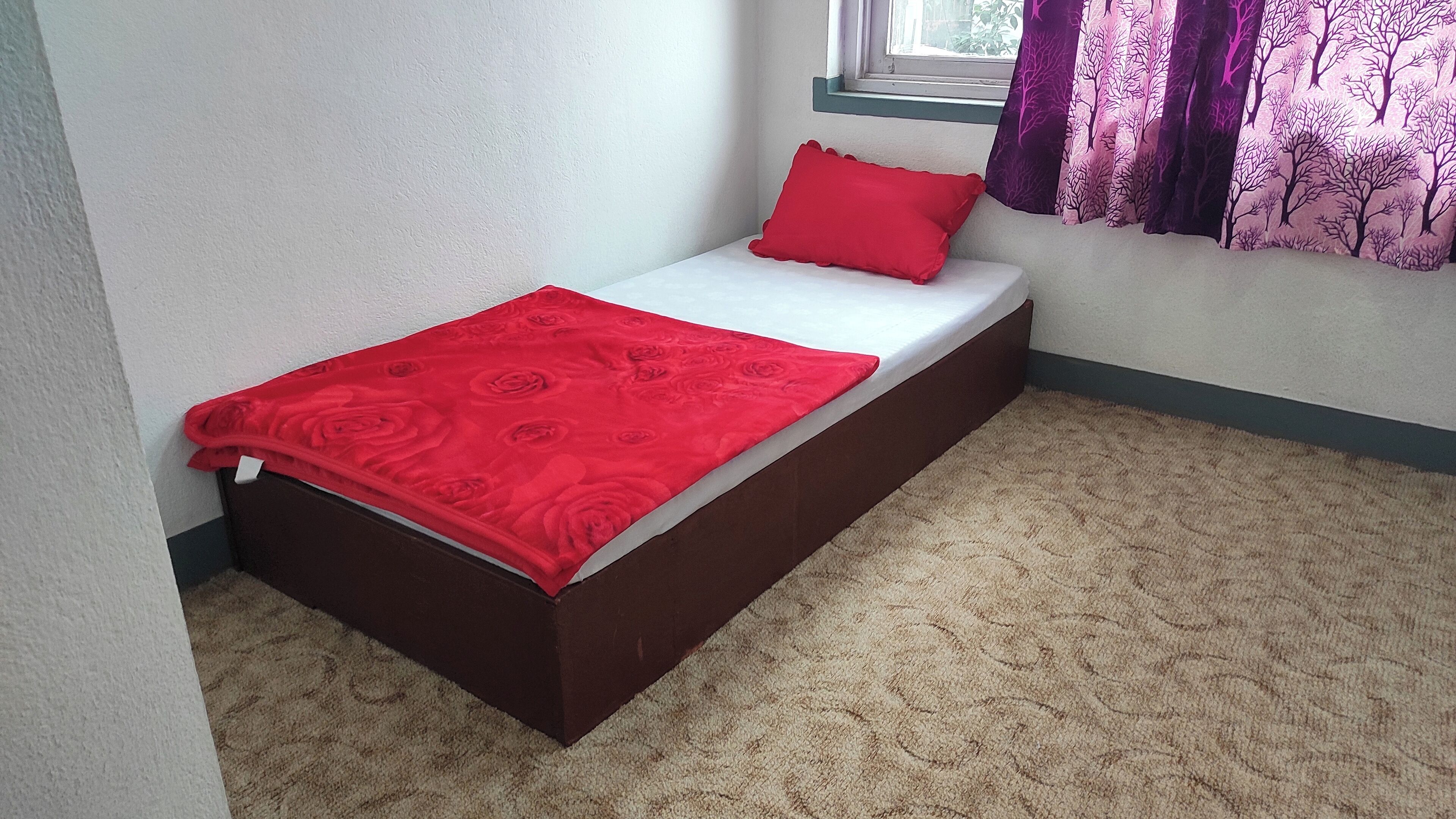 Een kleine eenpersoonskamer met een rood sprei, een passend rood kussen en een raam met paarse gordijnen.