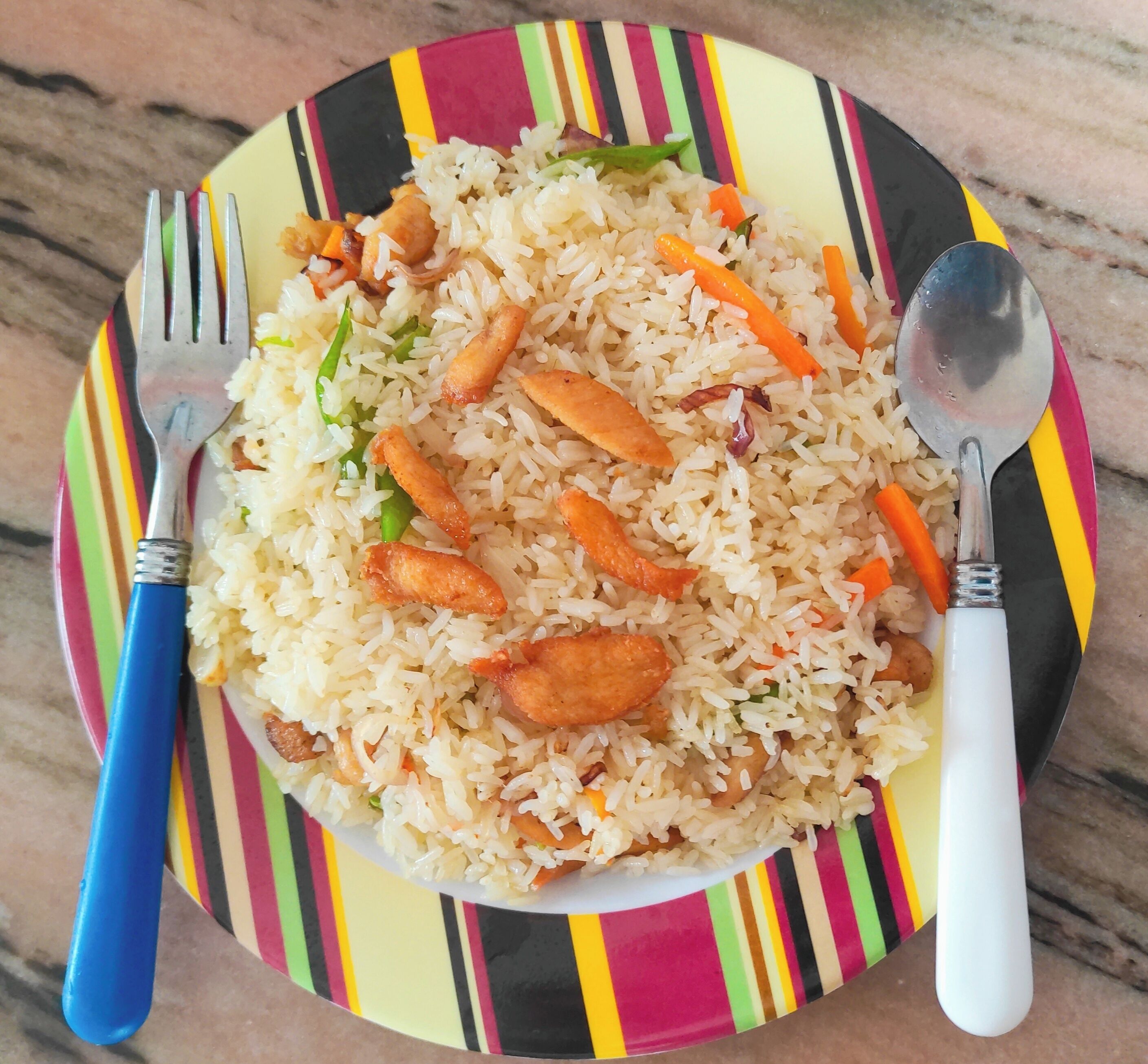 Une assiette de riz frit aux légumes et à la viande, servie dans une assiette colorée à rayures, accompagnée d’une fourchette et d’une cuillère.