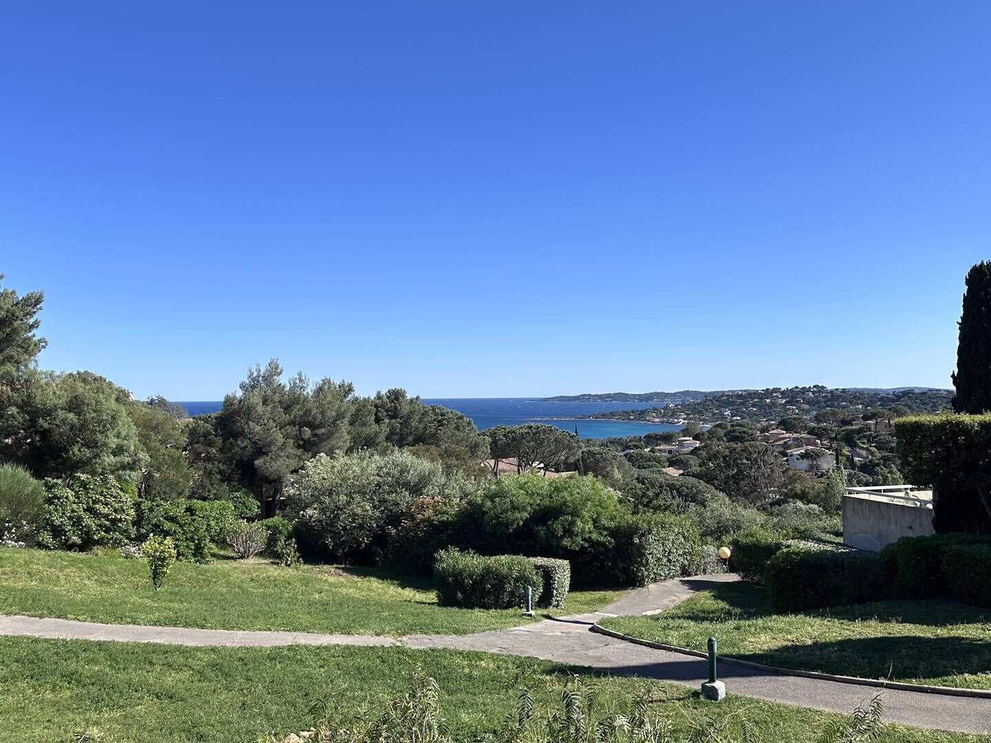 Uma área gramada com um caminho que leva a um edifício, com vista para uma cidade costeira e o mar.