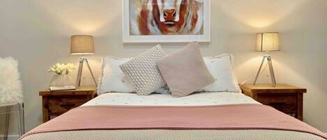 A bedroom with a bed, two bedside tables, two lamps, a painting on the wall, and a fluffy white pillow on the bed.