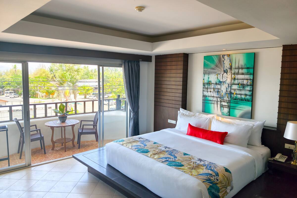 Een hotelkamer met een bed, een klein tafeltje met stoelen, een balkon met uitzicht op de straat en een schilderij aan de muur.