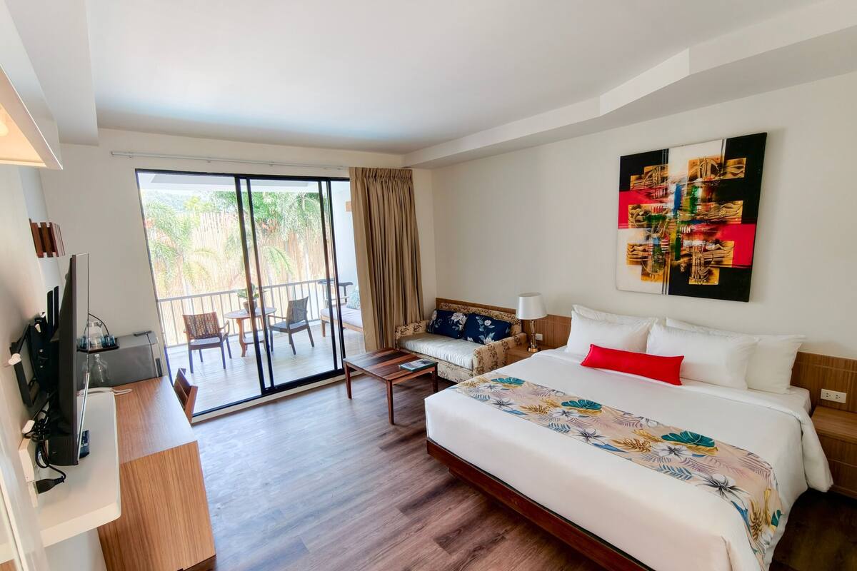 Een hotelkamer met een groot bed, een bureau met een computer, een balkon met stoelen en een schilderij aan de muur.