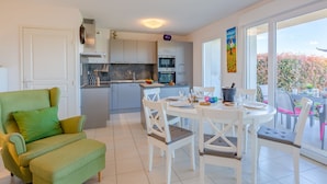 A modern kitchen with white cabinets, a dining area with white chairs, and a balcony with a view of trees.
