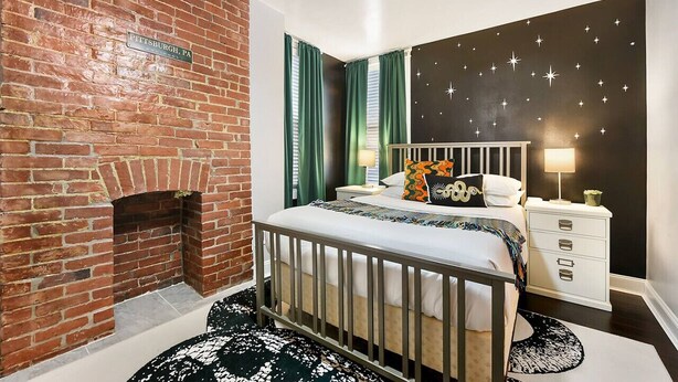 Een slaapkamer met een metselwerk open haard, een bed met een witte hoofdbord, groene gordijnen, een sterrenpatroon op de muur, en een nachtkastje met een lamp.