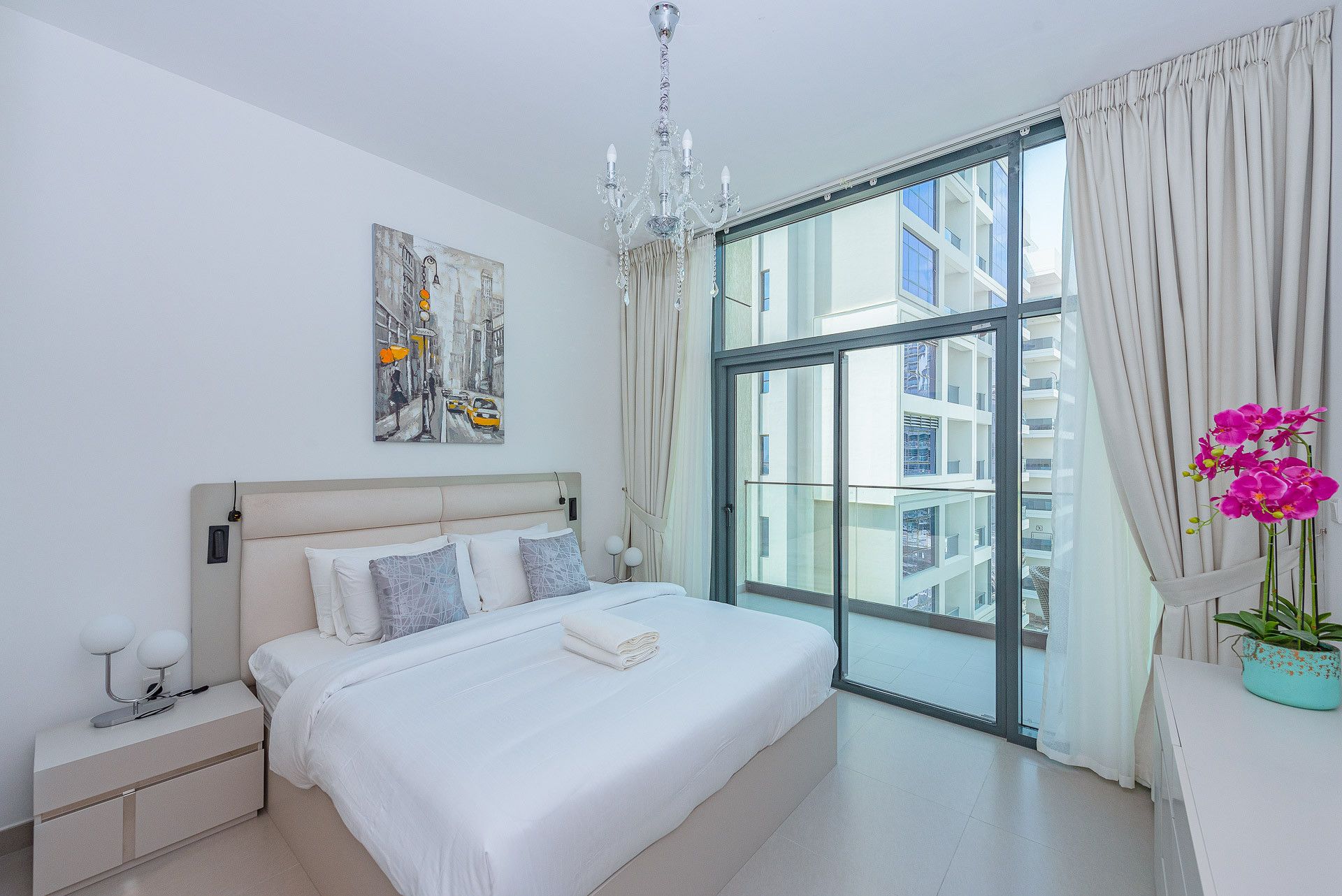 A modern bedroom with a large bed, a chandelier, a cityscape painting, and a view of buildings through a large window.