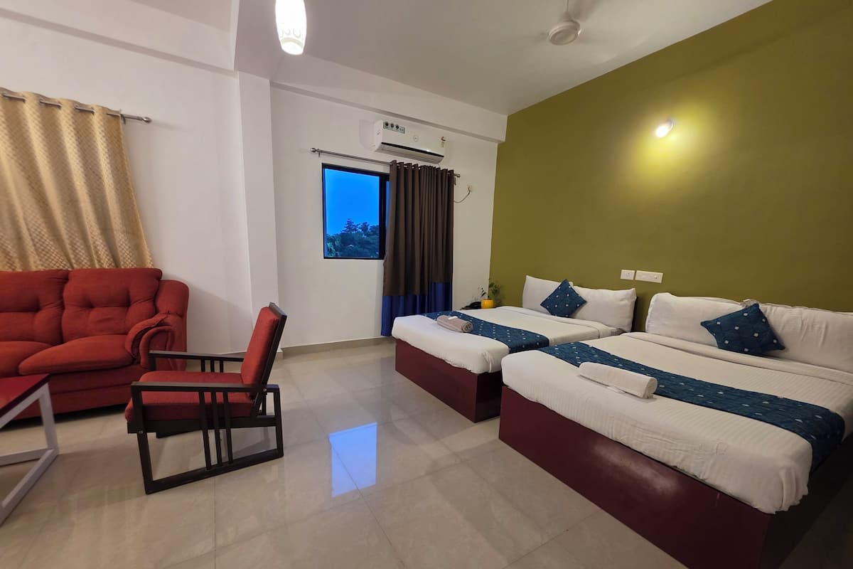 Habitación de hotel con sofá rojo, una silla, dos camas con ropa de cama blanca y azul, una ventana con vistas y un equipo de aire acondicionado.