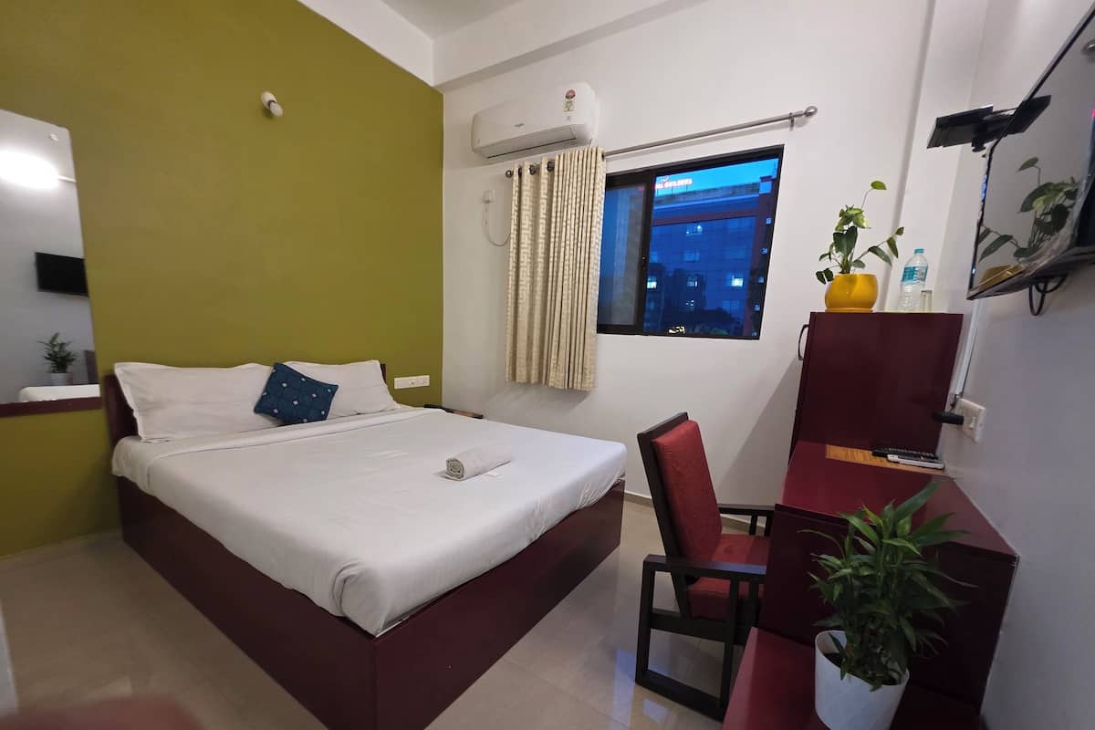 Habitación de hotel con cama, escritorio, silla, planta en maceta y ventana con vistas a edificios.