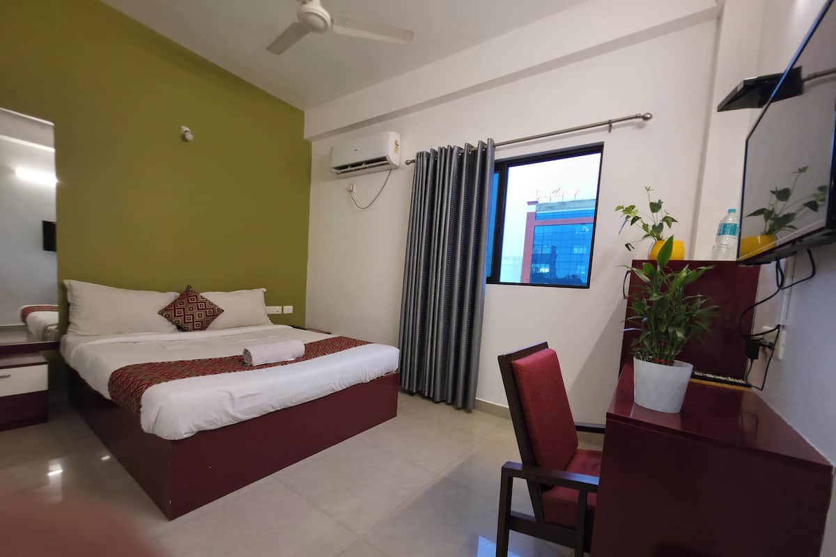 Habitación de hotel con cama, escritorio, silla, equipo de aire acondicionado, ventana con cortinas y una planta en maceta.