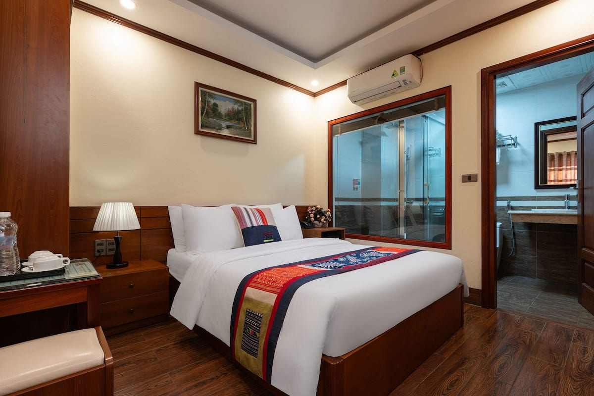 Habitación de hotel con una cama grande, una mesita de noche con lámpara, un escritorio de madera y una ventana con vistas al exterior.