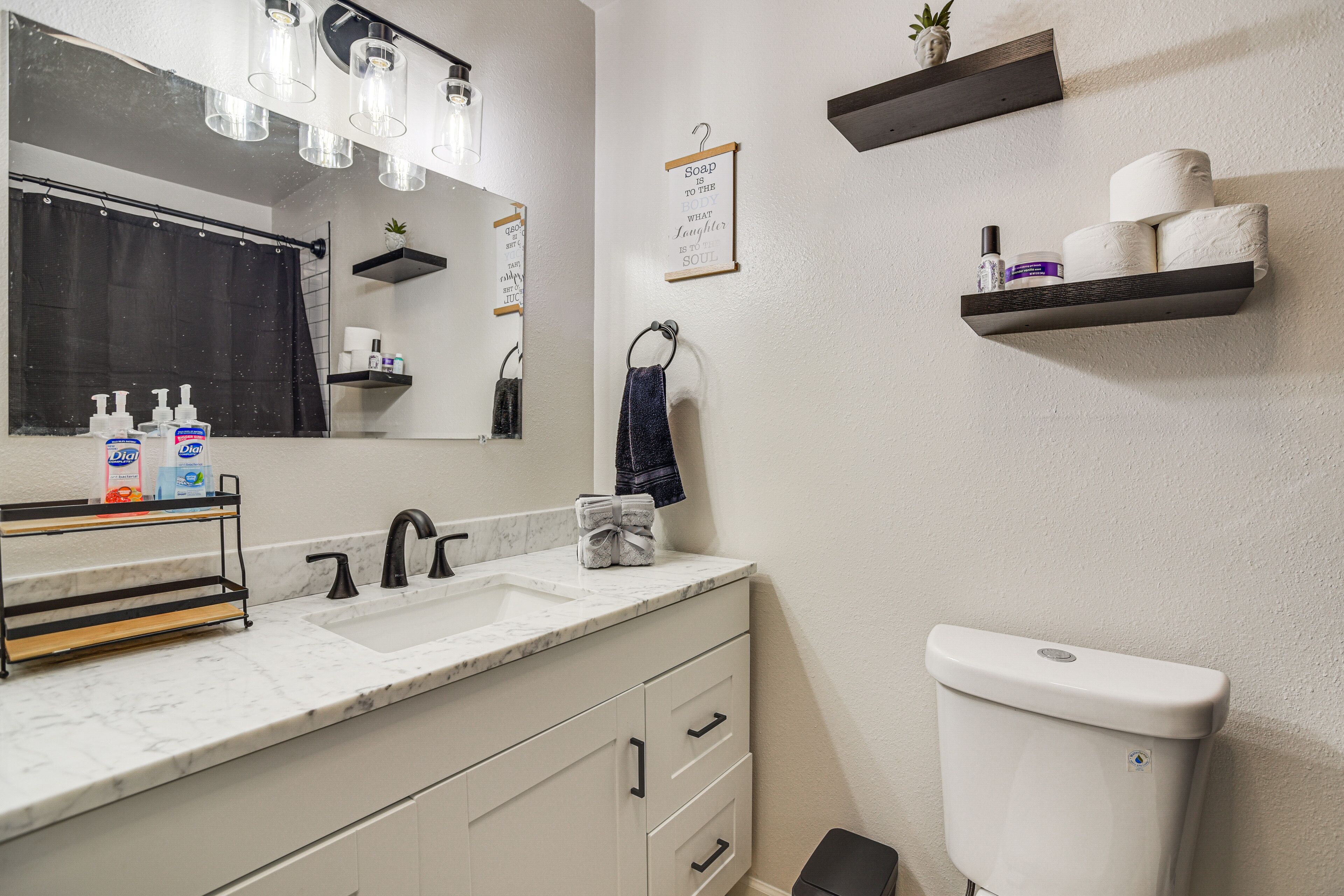 Ein Badezimmer mit einem weißen Waschbecken, einem schwarzen Wasserhahn und einer weißen Toilette. Es gibt Regale mit Toilettenartikeln und ein Handtuch, das an der Wand hängt.