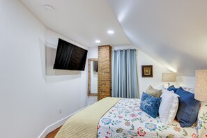 Un dormitorio con cama, televisión de pantalla plana en la pared, un pilar de ladrillo y ventana con cortinas.