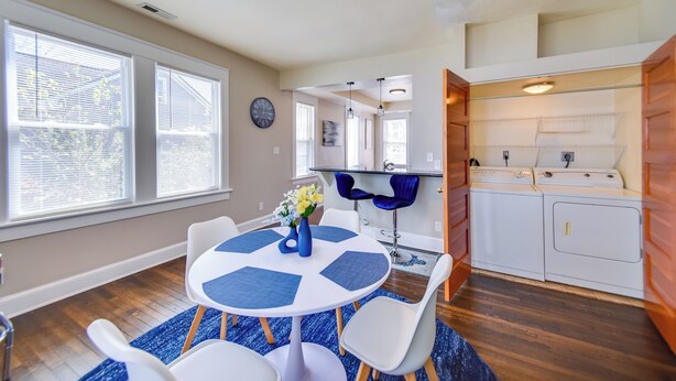 Een moderne keuken met witte apparaten, een eethoek met een ronde tafel en blauwe placemats, en een groot raam met zonwering.