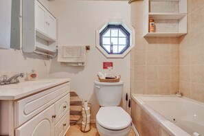 Et badeværelse med toilet, håndvask og badekar. Et vindue er placeret over toilettet, et håndklæde hænger på væggen, og der er en hylde med toiletartikler.