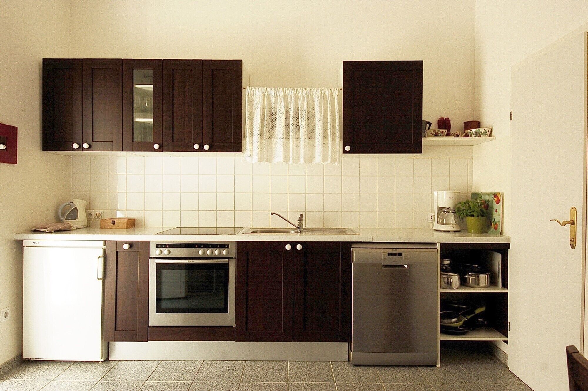 Een kleine keuken met donkere kastjes, een witte betegelde spatwand, een kookplaat, een oven, een vaatwasser en een koffiezetapparaat.
