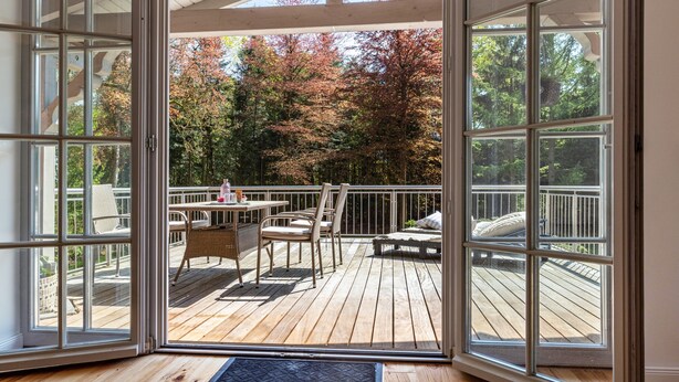 Een open glazen deur die leidt naar een terras met een tafel en stoelen, omgeven door bomen.