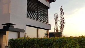 Um edifício moderno com uma grande janela, um telhado plano e uma sebe em primeiro plano.
