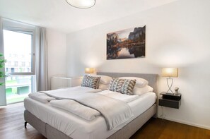 Une chambre moderne avec un grand lit, deux tables de chevet, une lampe fixée au mur et une fenêtre donnant sur des immeubles.