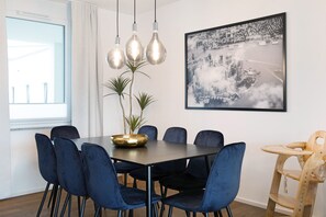 Un coin repas avec une table sombre, des chaises bleues, un centre de table en forme de bol doré, des luminaires suspendus et une œuvre d’art représentant un paysage urbain accrochée au mur.