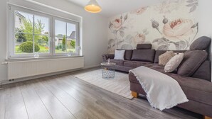 A living room with a large window, a sofa, a side table, and a floral wallpaper. - Inn 'Haus Am Garten' with Private Terrace, Garden and Balcony (Alfeld (Leine))