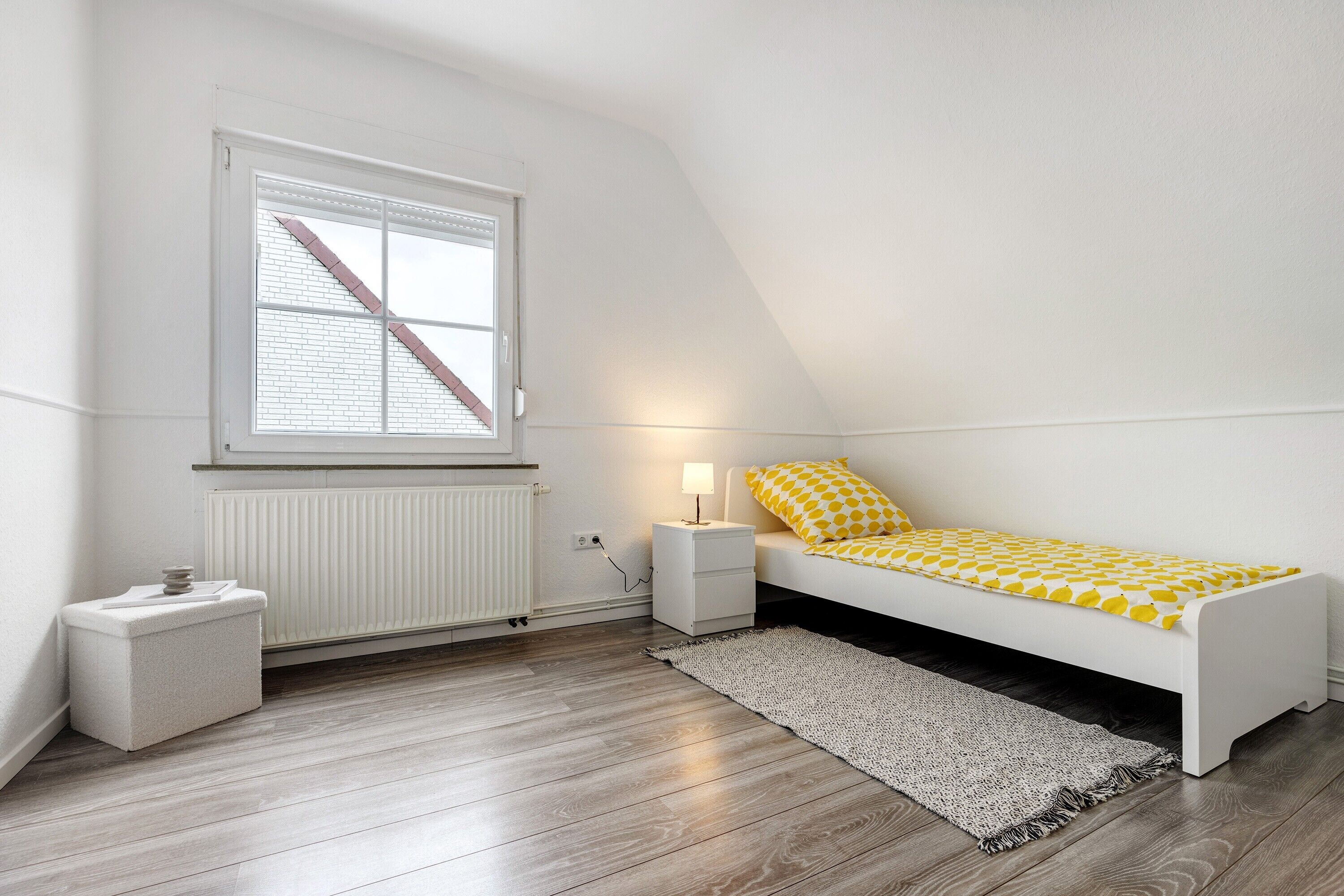 A bedroom with a white bed, a nightstand, a lamp, a radiator, a window with blinds, and a small white storage unit.
