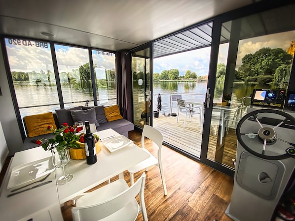 A modern interior with a dining area, sofa, and large windows overlooking a body of water.