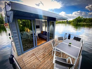 A modern houseboat with a covered deck, glass doors, and a dining area.
