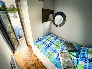 A small, compact room with a bed, a round window, and a view of a body of water outside.