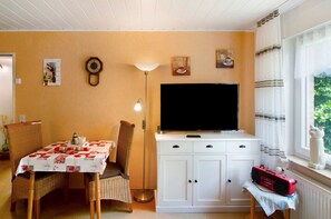 A room with a television, a cabinet, a table with a floral tablecloth, a chair, a lamp, and a window with curtains. - Apartment 'Ferienhaus Gerda Wohnung 1' with Private Terrace, Private Garden and Wi-Fi (Ochtersum)