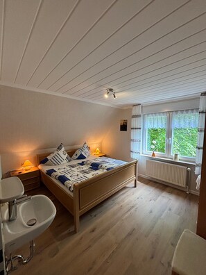 A bedroom with a wooden bed, bedside lamps, a window with curtains, a radiator, and a bathroom with a sink. - Apartment 'Ferienhaus Gerda Wohnung 1' with Private Terrace, Private Garden and Wi-Fi (Ochtersum)