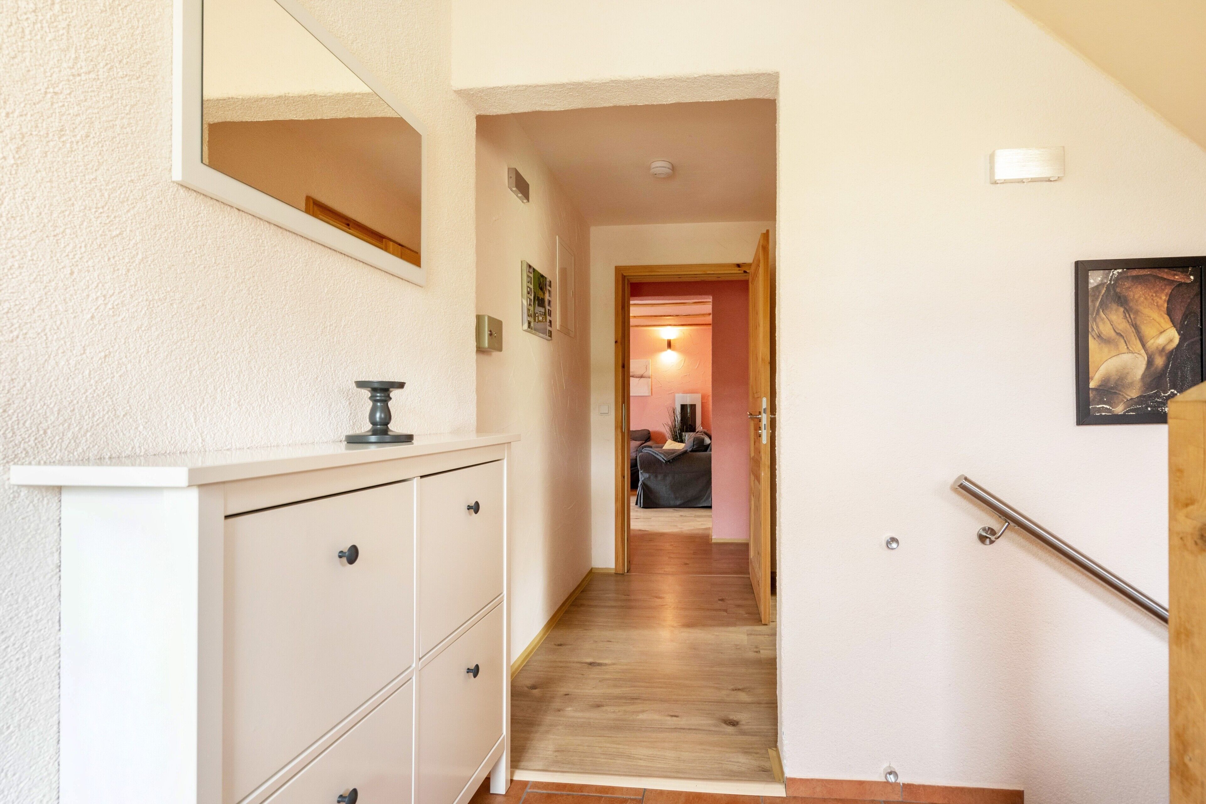 Un couloir avec un plancher en bois, une commode blanche, un miroir et une porte qui donne sur une pièce contenant un lit et une lampe.