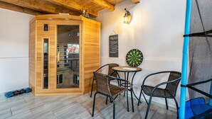A sauna room with wooden walls and a glass door, a dartboard on the wall, and two wicker chairs around a small table. - Holiday Home 'Ferienhaus Fichtelberg' with Mountain View, Private Terrace and Wi-Fi (Oberwiesenthal)