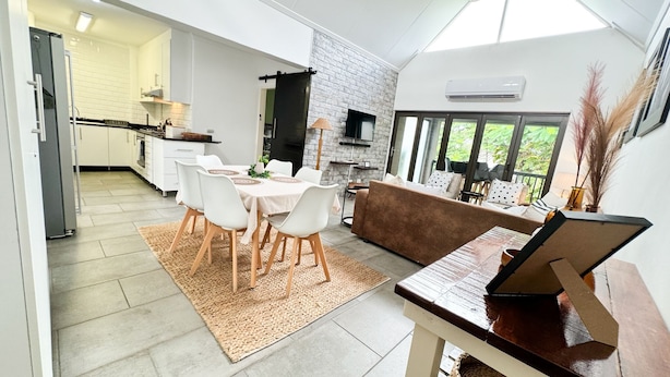 A modern kitchen with white cabinets, a dining area with a white table and chairs, a brown sofa, and a large window with a view of greenery. - Luxury Apartment St Lucia 43 On the Bridge (Saint Lucia)
