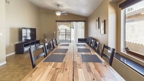 A dining area with a long wooden table, black chairs, a ceiling fan, and a TV on a stand.