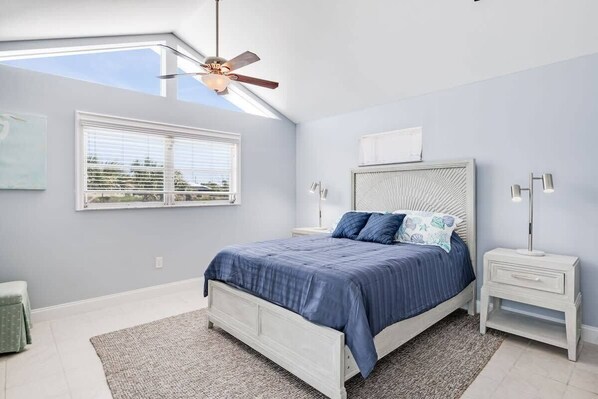 Ein Schlafzimmer mit Dachfenster, einem Bett mit blauer Bettwäsche, einem weißen Nachttisch mit Lampe und einem Deckenventilator.
