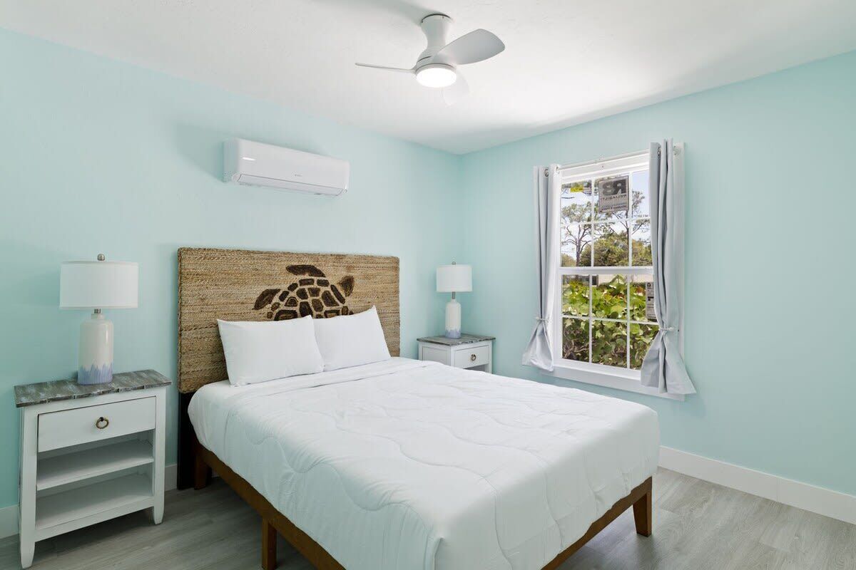 Quarto com cama, duas mesas de cabeceira, ventoinha no teto, aparelho de ar condicionado, janela com cortinas e uma obra de arte na parede.