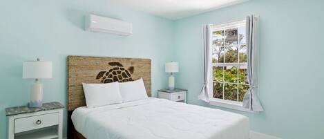 Quarto com cama, duas mesas de cabeceira, ventoinha no teto, aparelho de ar condicionado, janela com cortinas e uma obra de arte na parede.