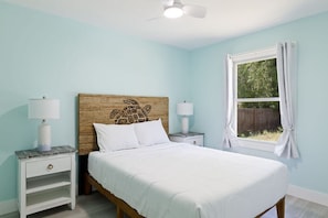 Un dormitorio con cama, dos mesitas de noche, ventilador de techo, ventana con cortinas y vistas al exterior.