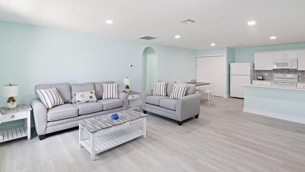 Ett modernt vardagsrum med två soffor, ett soffbord och en köksdel med vita skåp och vitvaror.